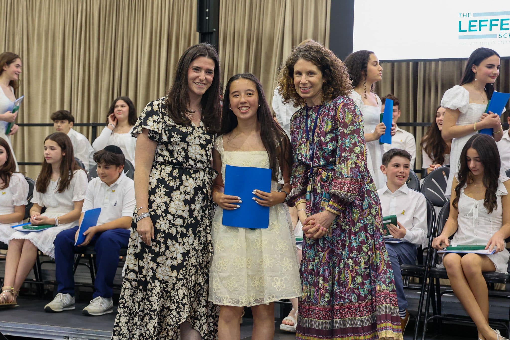 A group of three people, one in the middle and two on each side, are smiling for camera. They are at a ceremony, as shown by the backdrop displaying "The Leffell School." The central figure, a younger student in white clothes , on the sides are two women in floral dresses.