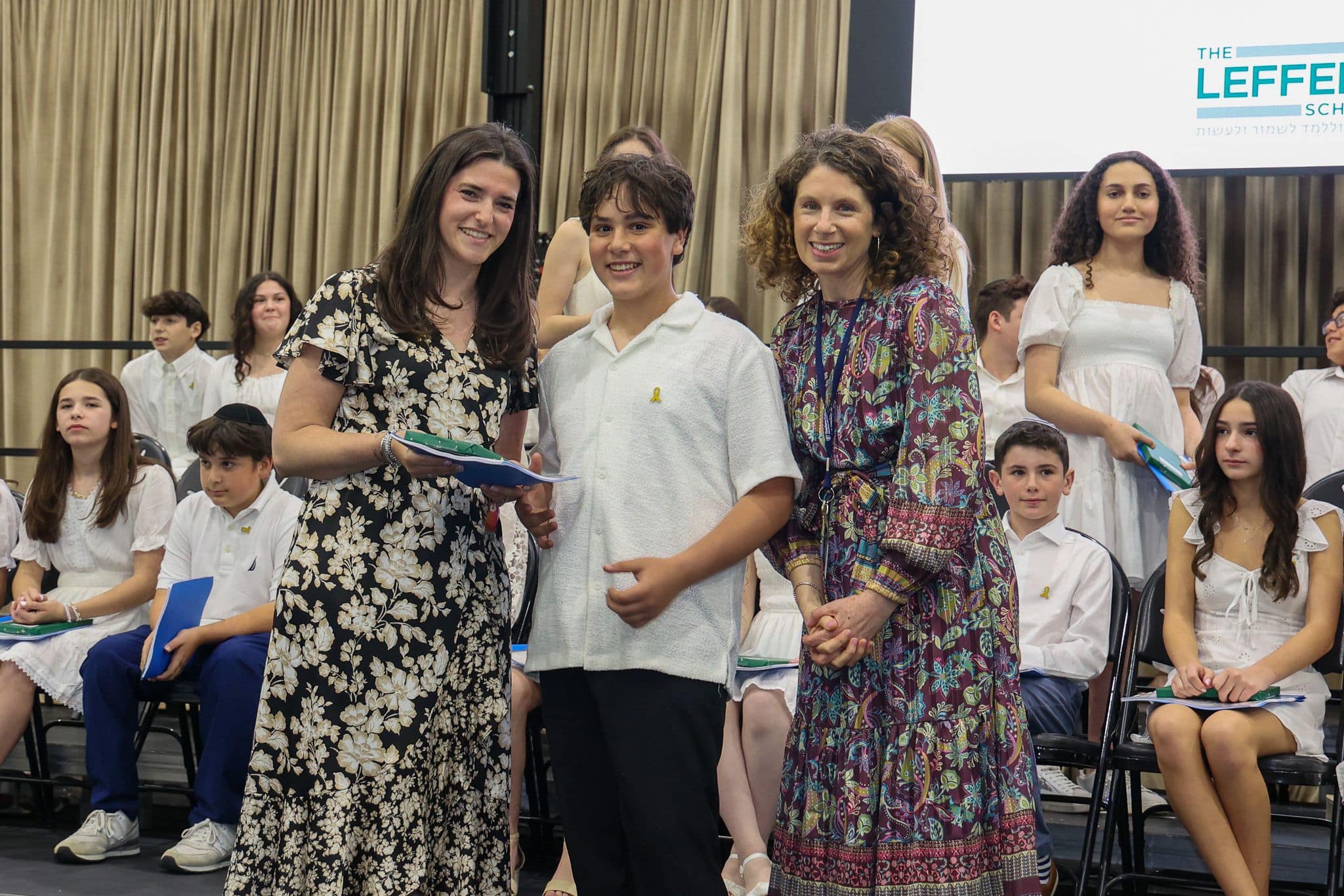 A group of three people, one in the middle and two on each side, are smiling for camera. They are at a ceremony, as shown by the backdrop displaying "The Leffell School." The central figure, a younger student in white clothes , on the sides are two women in floral dresses.
