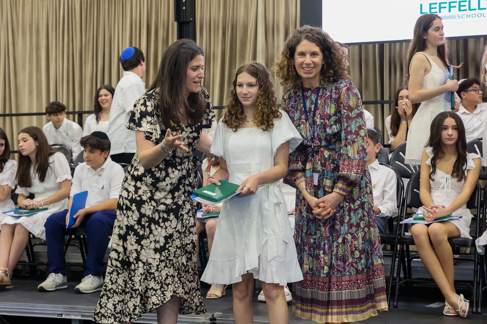 A group of three people, one in the middle and two on each side, are smiling for camera. They are at a ceremony, as shown by the backdrop displaying "The Leffell School." The central figure, a younger student in white clothes , on the sides are two women in floral dresses.