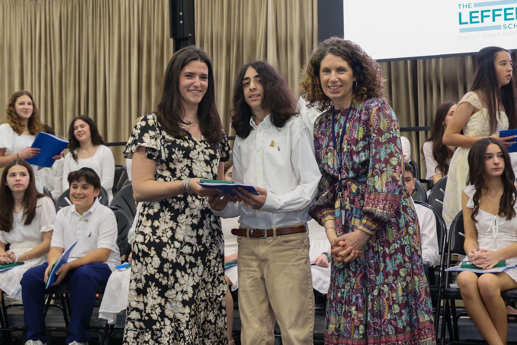 A group of three people, one in the middle and two on each side, are smiling for camera. They are at a ceremony, as shown by the backdrop displaying "The Leffell School." The central figure, a younger student in white clothes , on the sides are two women in floral dresses.