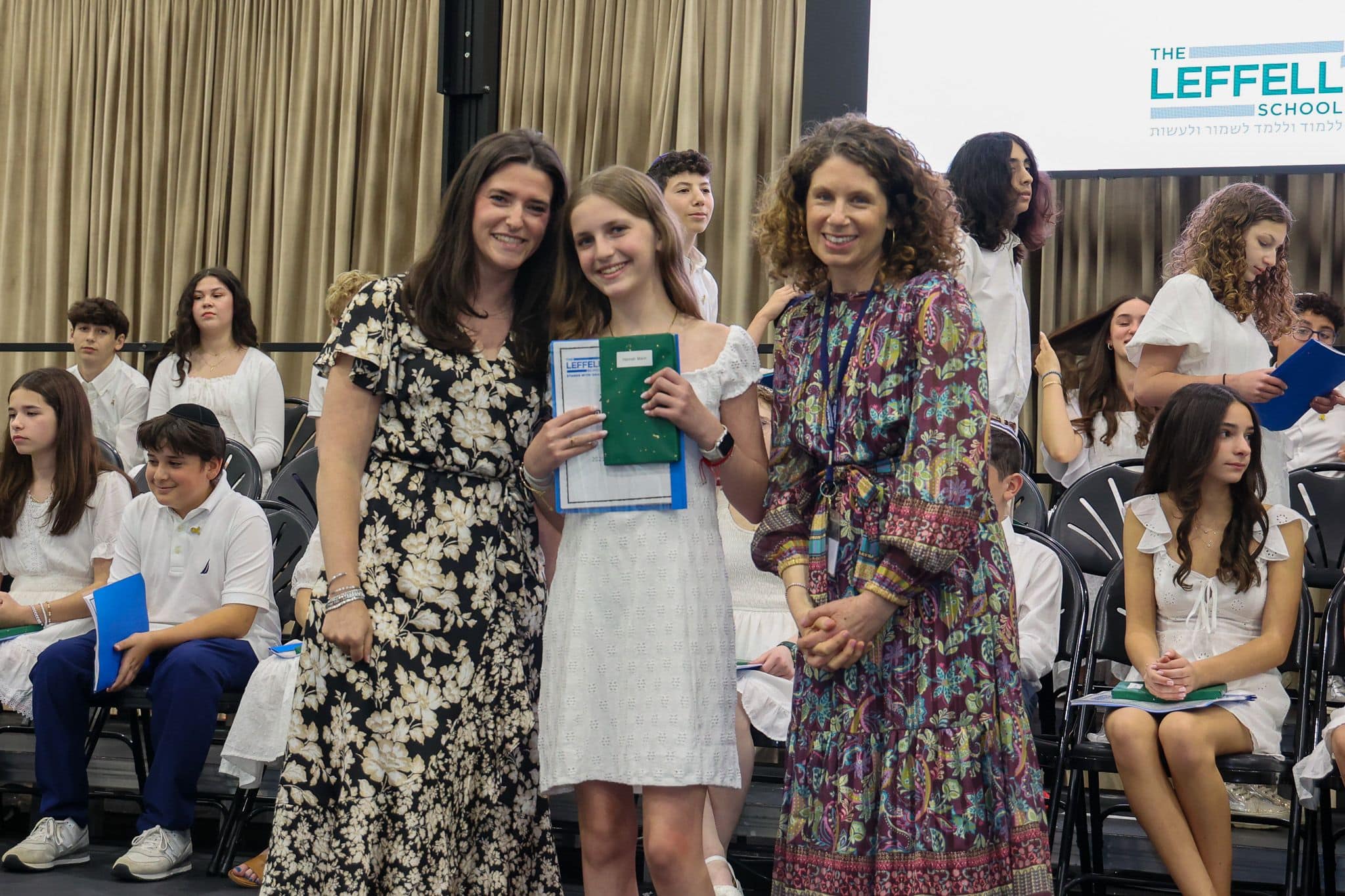 A group of three people, one in the middle and two on each side, are smiling for camera. They are at a ceremony, as shown by the backdrop displaying "The Leffell School." The central figure, a younger student in white clothes , on the sides are two women in floral dresses.