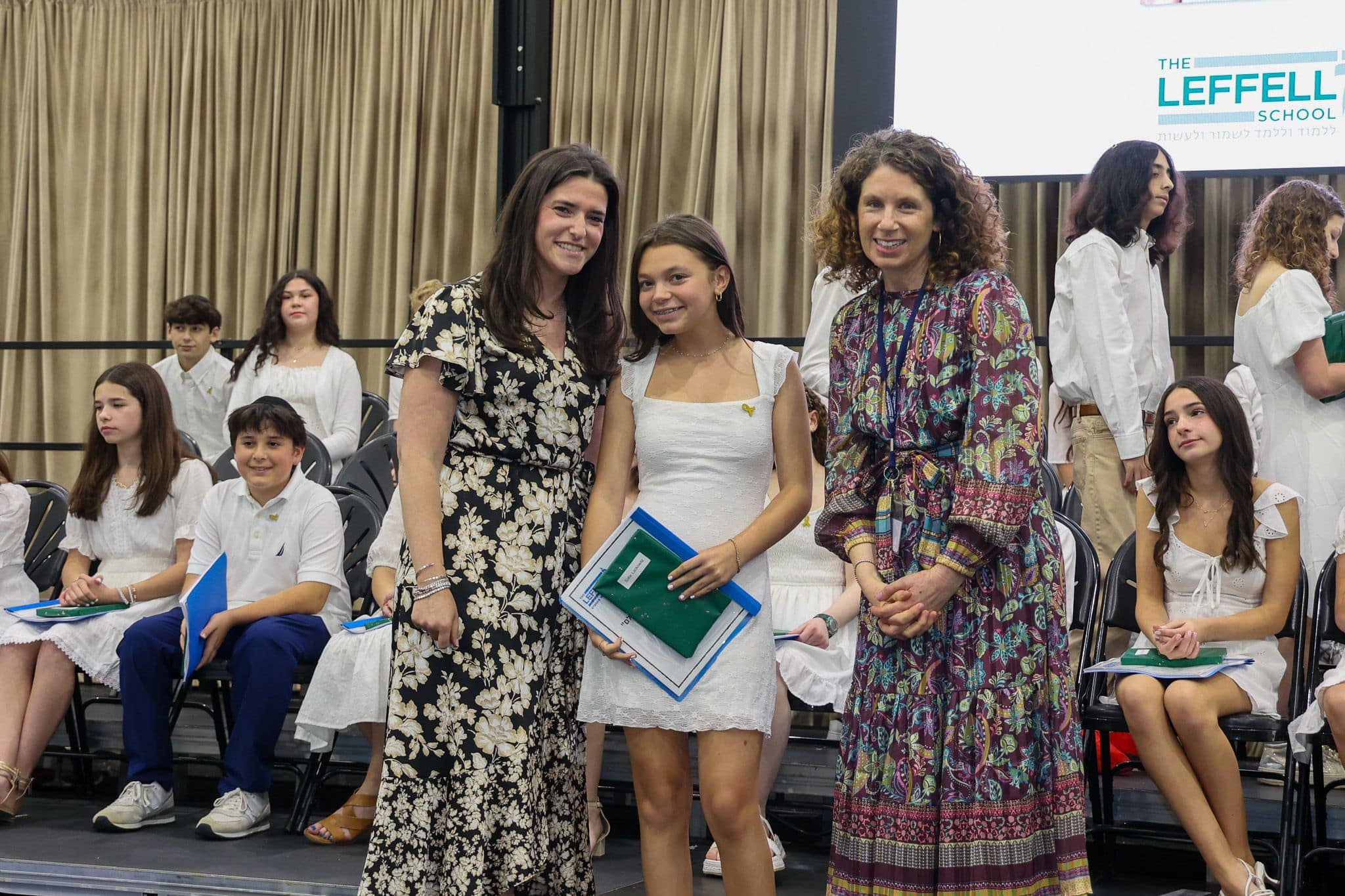 A group of three people, one in the middle and two on each side, are smiling for camera. They are at a ceremony, as shown by the backdrop displaying "The Leffell School." The central figure, a younger student in white clothes , on the sides are two women in floral dresses.