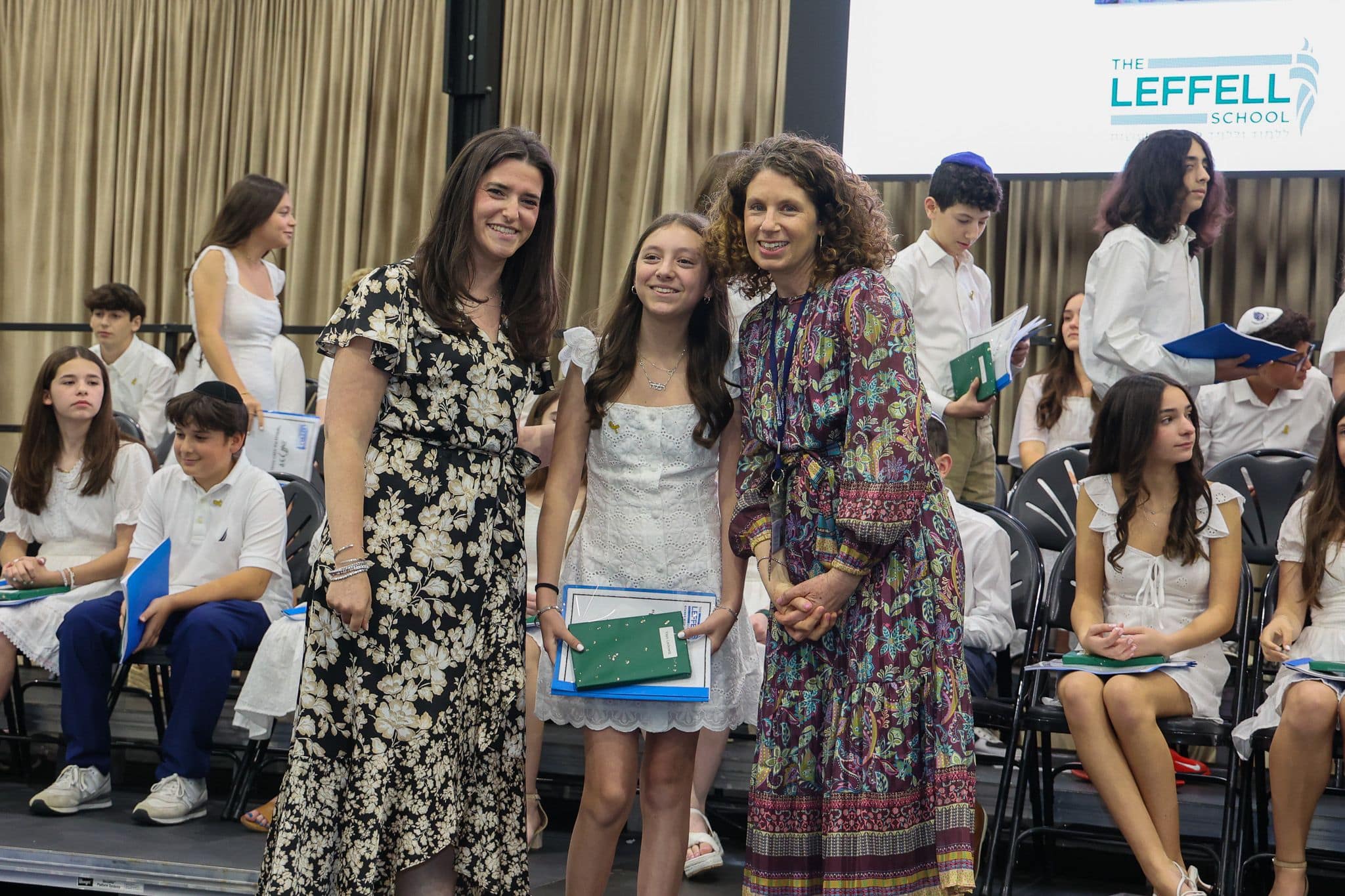 A group of three people, one in the middle and two on each side, are smiling for camera. They are at a ceremony, as shown by the backdrop displaying "The Leffell School." The central figure, a younger student in white clothes , on the sides are two women in floral dresses.