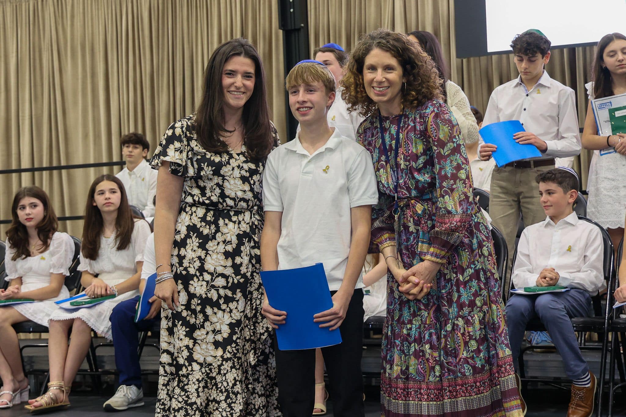 A group of three people, one in the middle and two on each side, are smiling for camera. They are at a ceremony, as shown by the backdrop displaying "The Leffell School." The central figure, a younger student in white clothes , on the sides are two women in floral dresses.