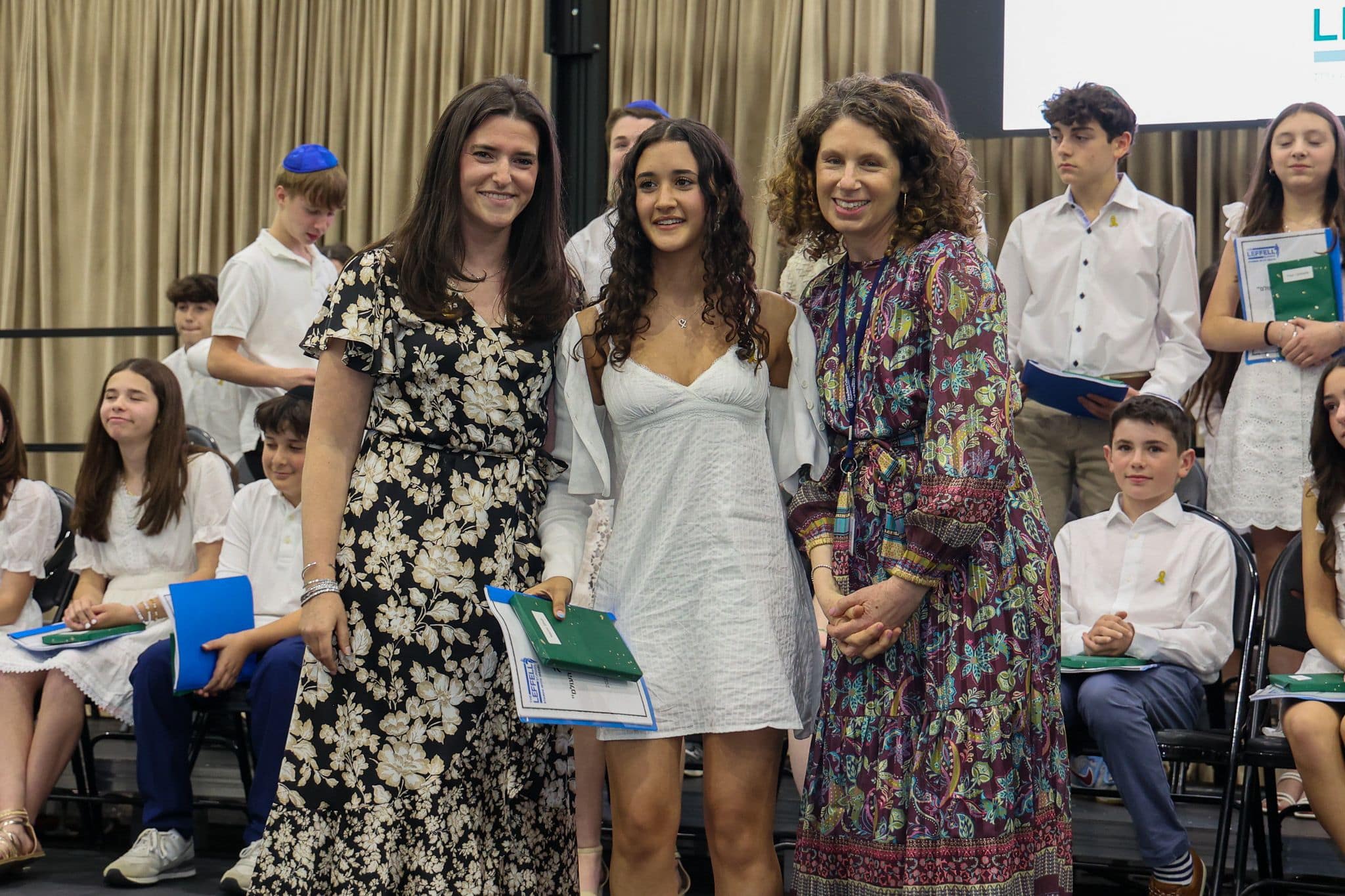 A group of three people, one in the middle and two on each side, are smiling for camera. They are at a ceremony, as shown by the backdrop displaying "The Leffell School." The central figure, a younger student in white clothes , on the sides are two women in floral dresses.