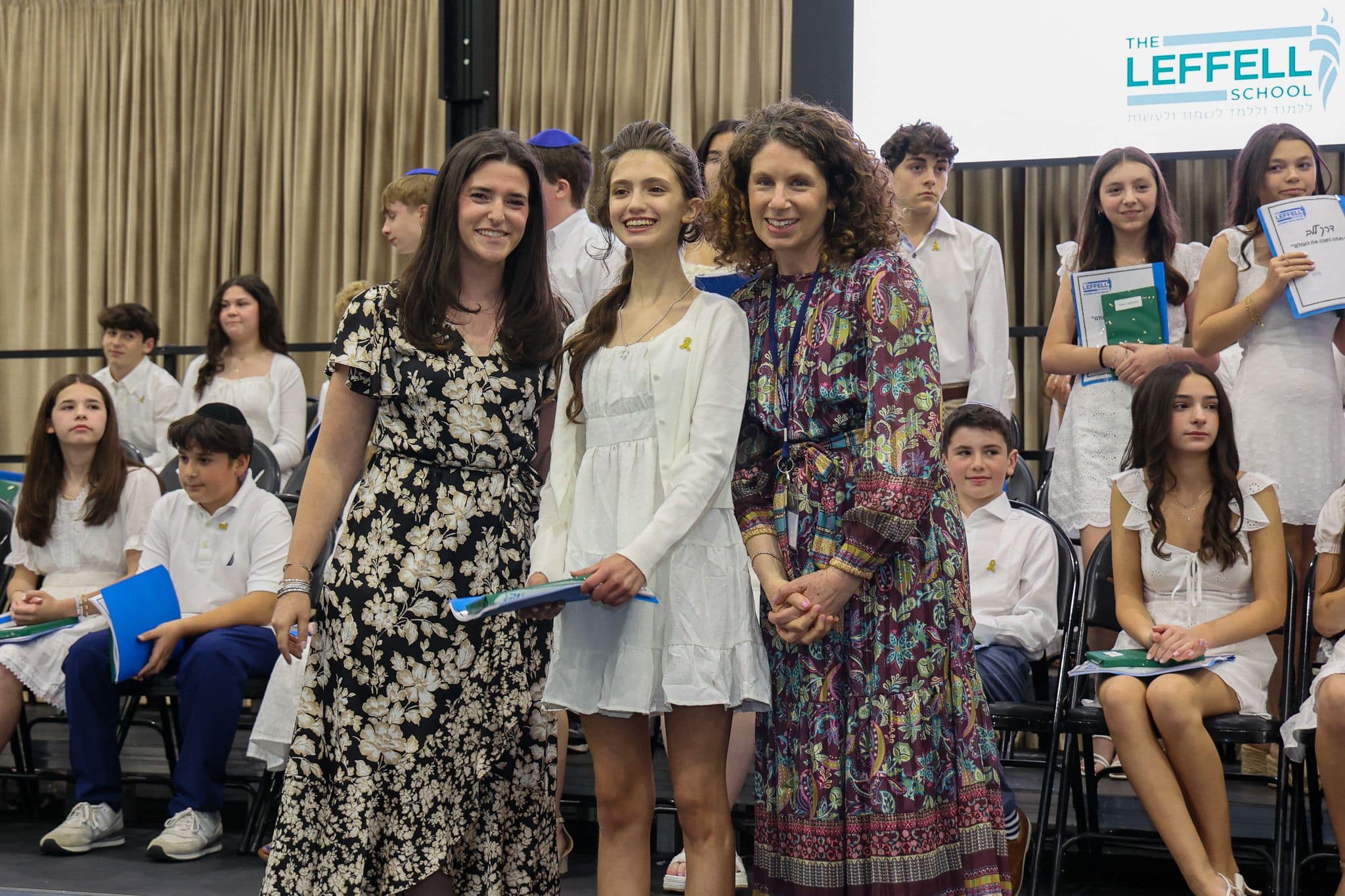A group of three people, one in the middle and two on each side, are smiling for camera. They are at a ceremony, as shown by the backdrop displaying "The Leffell School." The central figure, a younger student in white clothes , on the sides are two women in floral dresses.