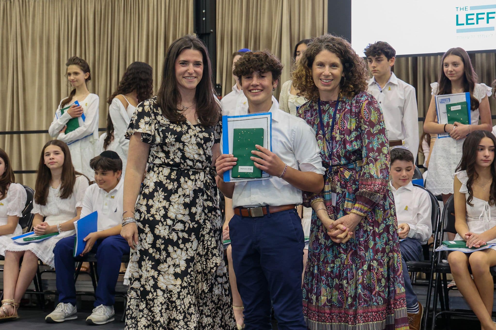 A group of three people, one in the middle and two on each side, are smiling for camera. They are at a ceremony, as shown by the backdrop displaying "The Leffell School." The central figure, a younger student in white clothes , on the sides are two women in floral dresses.