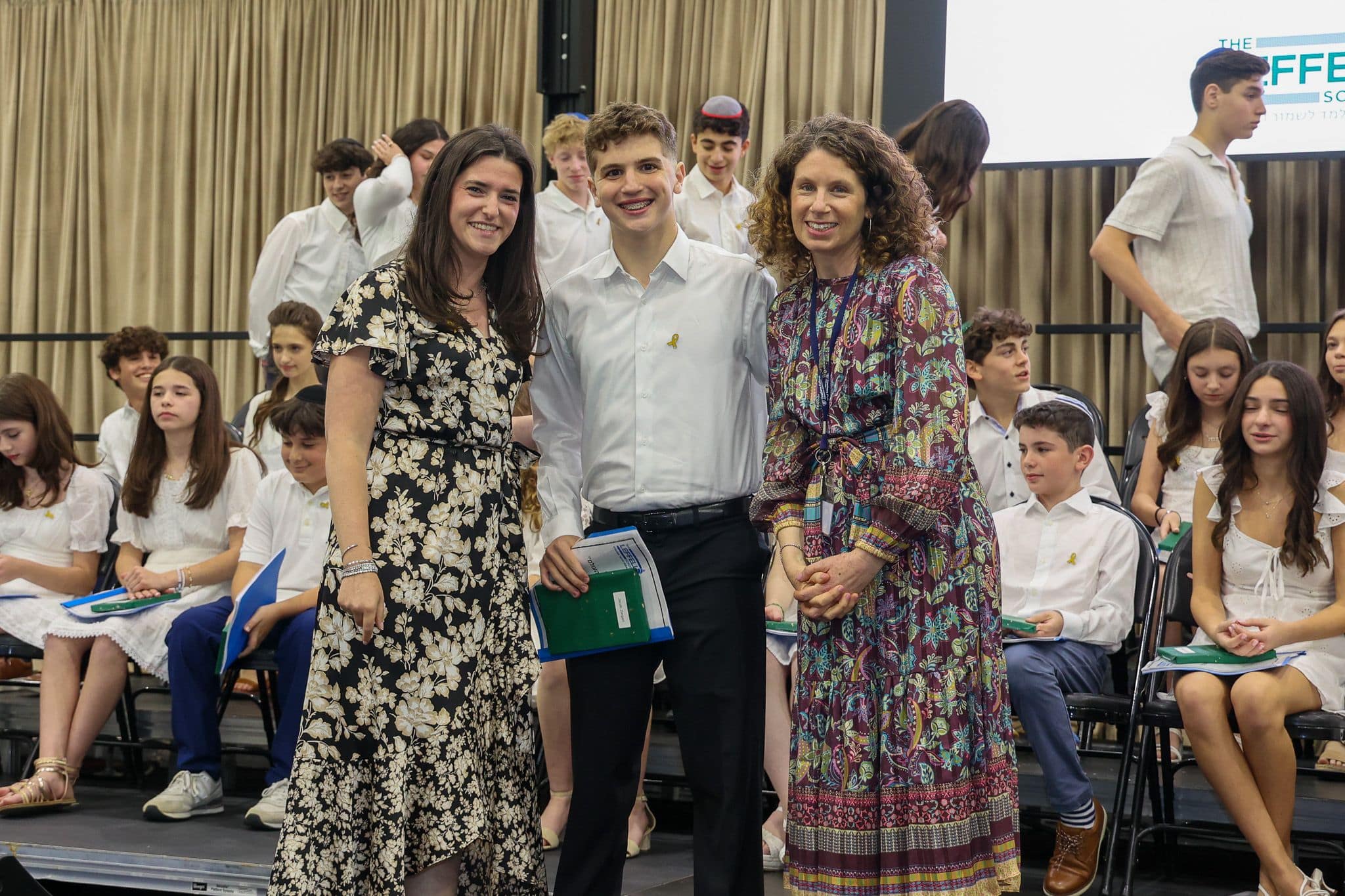 A group of three people, one in the middle and two on each side, are smiling for camera. They are at a ceremony, as shown by the backdrop displaying "The Leffell School." The central figure, a younger student in white clothes , on the sides are two women in floral dresses.