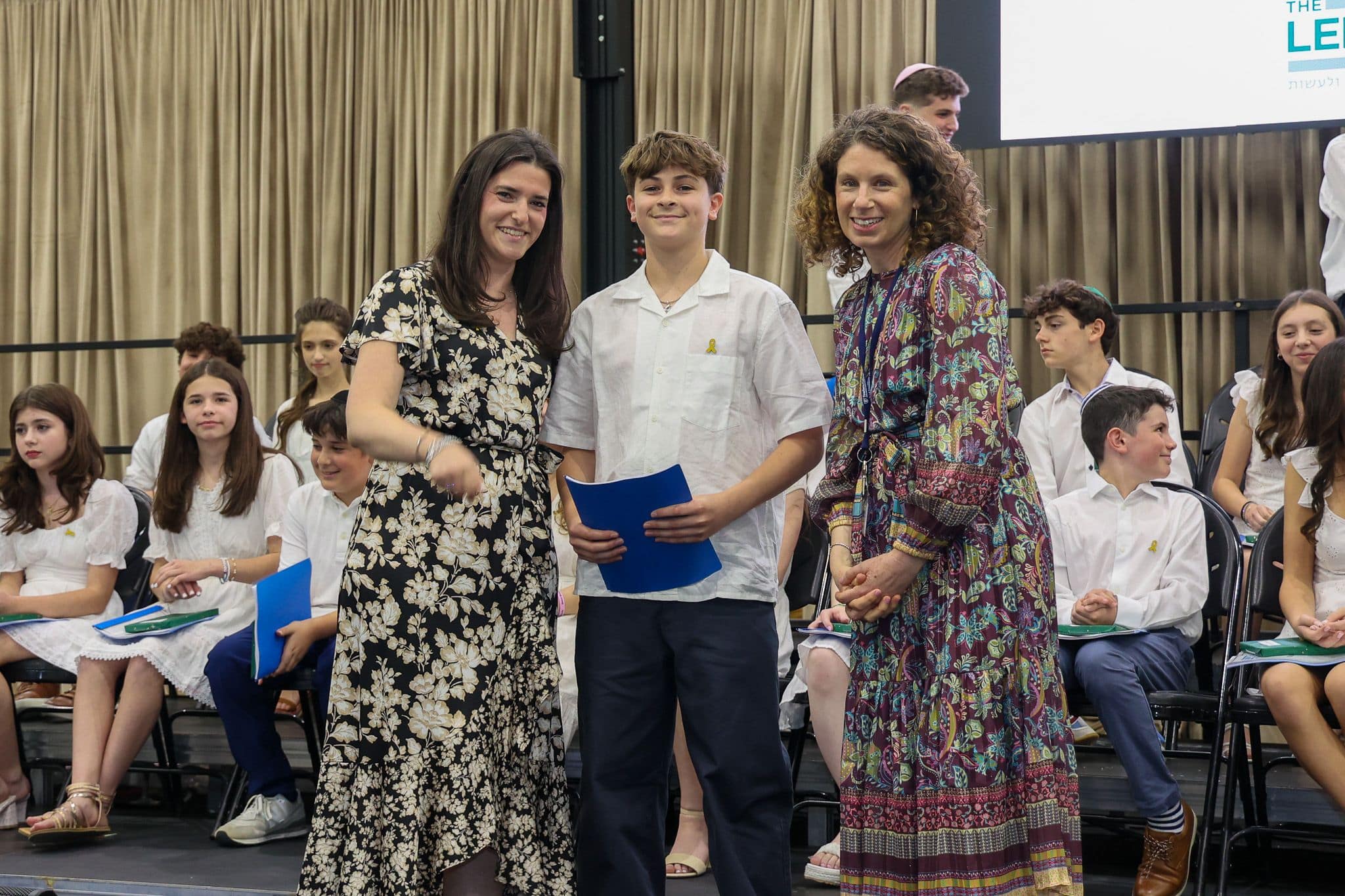 A group of three people, one in the middle and two on each side, are smiling for camera. They are at a ceremony, as shown by the backdrop displaying "The Leffell School." The central figure, a younger student in white clothes , on the sides are two women in floral dresses.