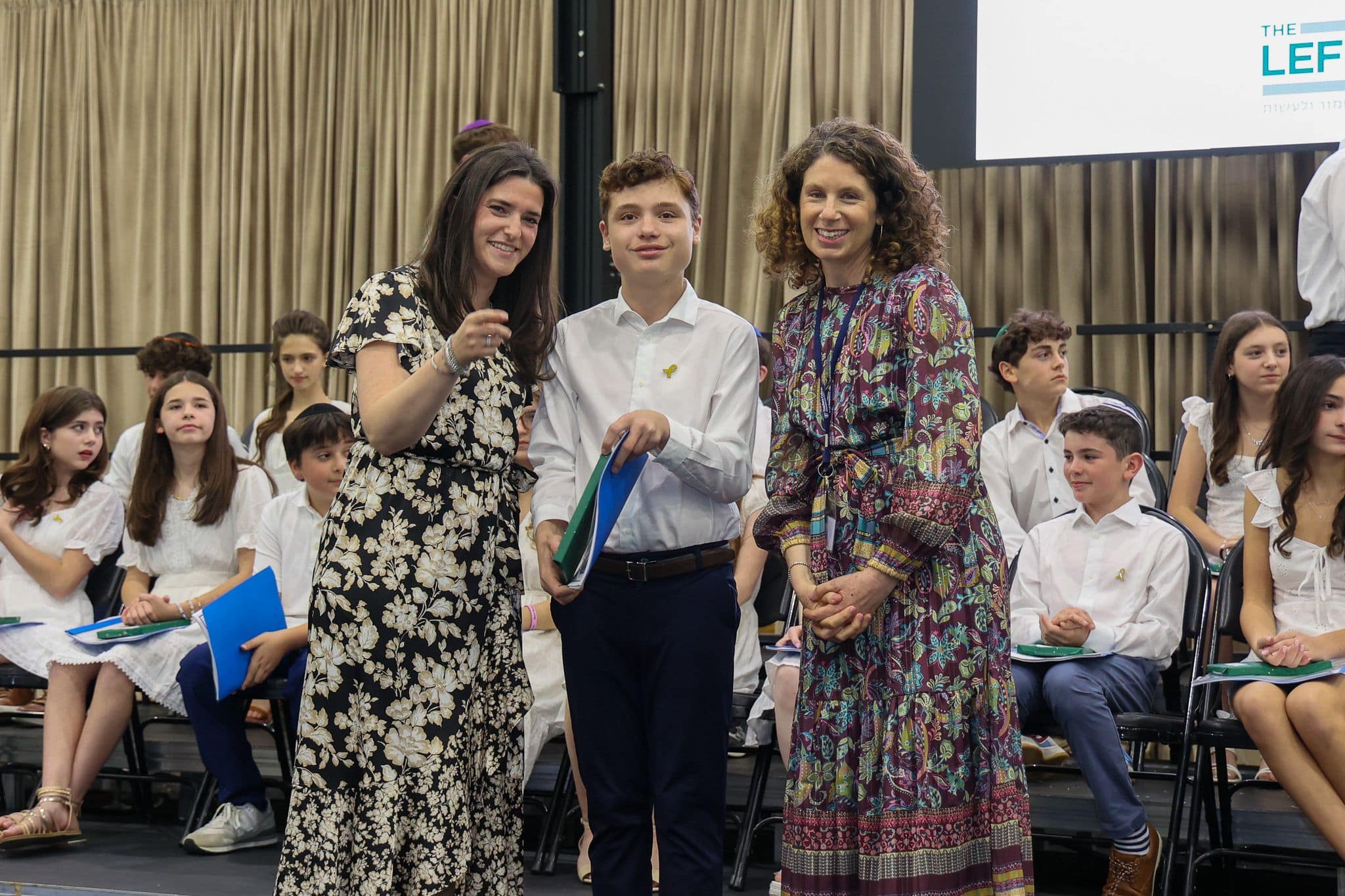 A group of three people, one in the middle and two on each side, are smiling for camera. They are at a ceremony, as shown by the backdrop displaying "The Leffell School." The central figure, a younger student in white clothes , on the sides are two women in floral dresses.