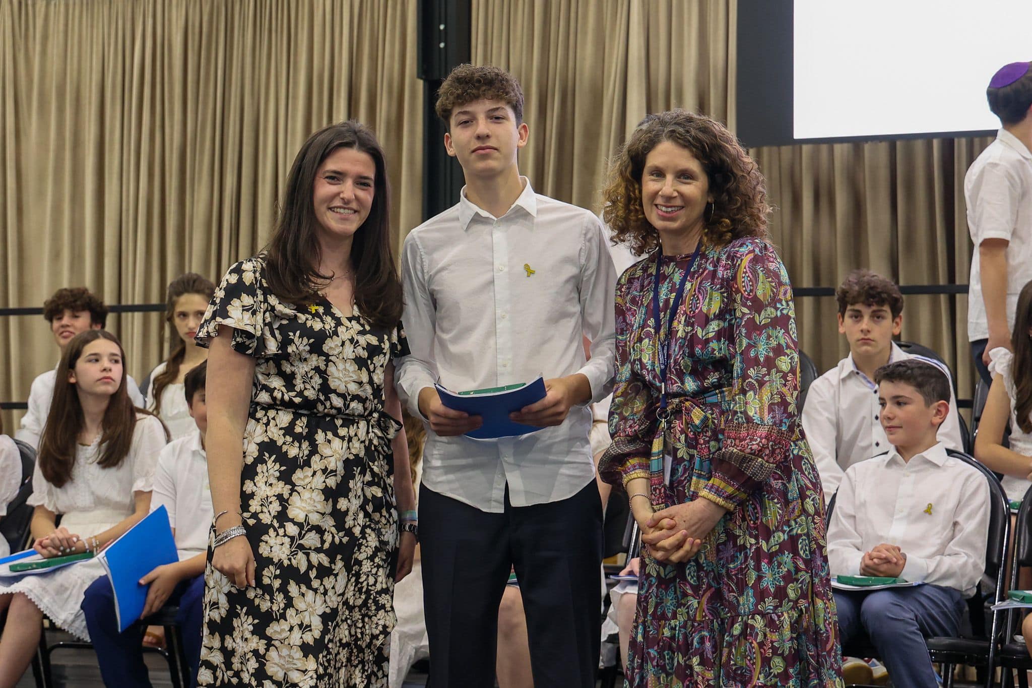 A group of three people, one in the middle and two on each side, are smiling for camera. They are at a ceremony, as shown by the backdrop displaying "The Leffell School." The central figure, a younger student in white clothes , on the sides are two women in floral dresses.