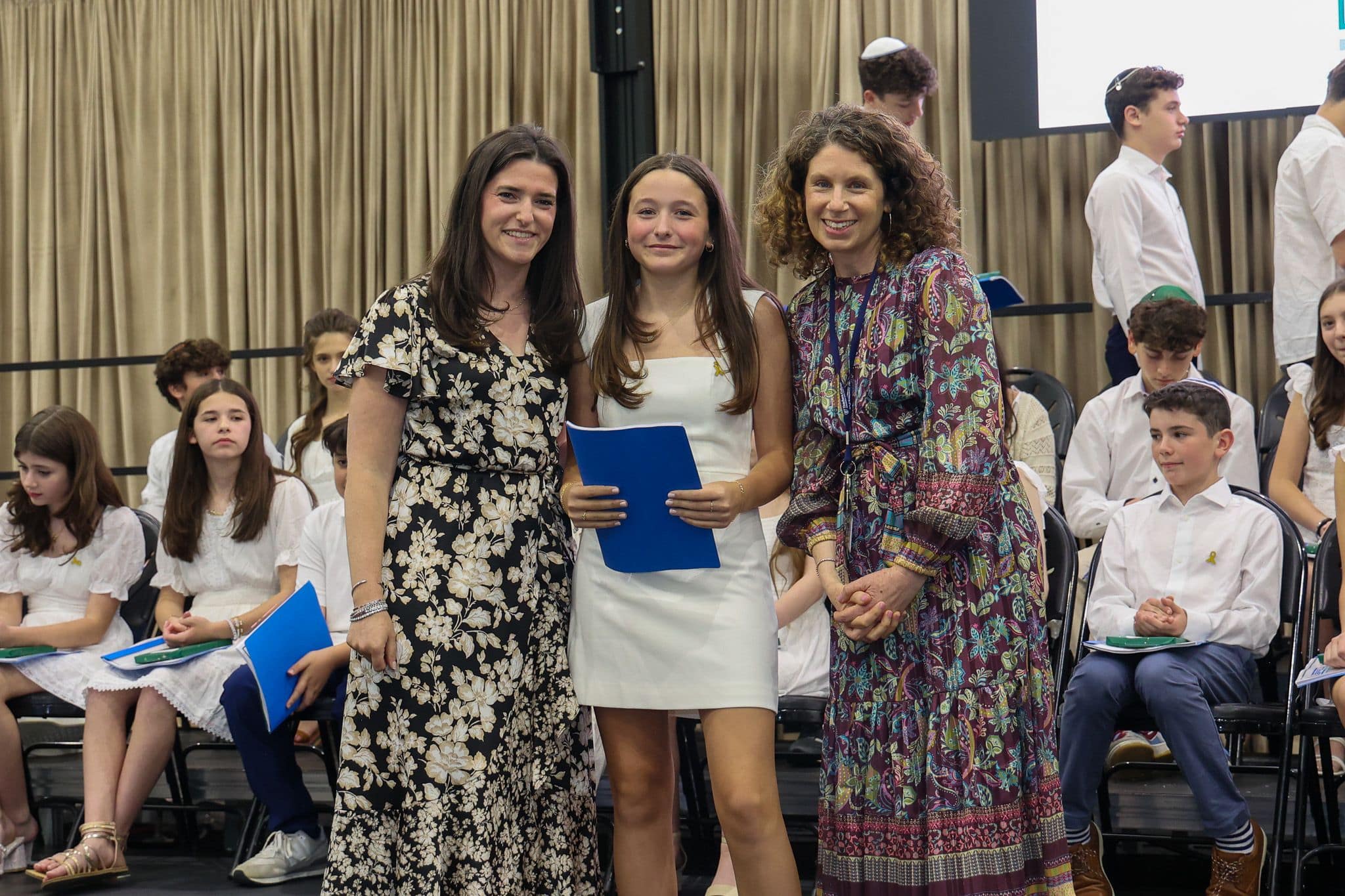 A group of three people, one in the middle and two on each side, are smiling for camera. They are at a ceremony, as shown by the backdrop displaying "The Leffell School." The central figure, a younger student in white clothes , on the sides are two women in floral dresses.