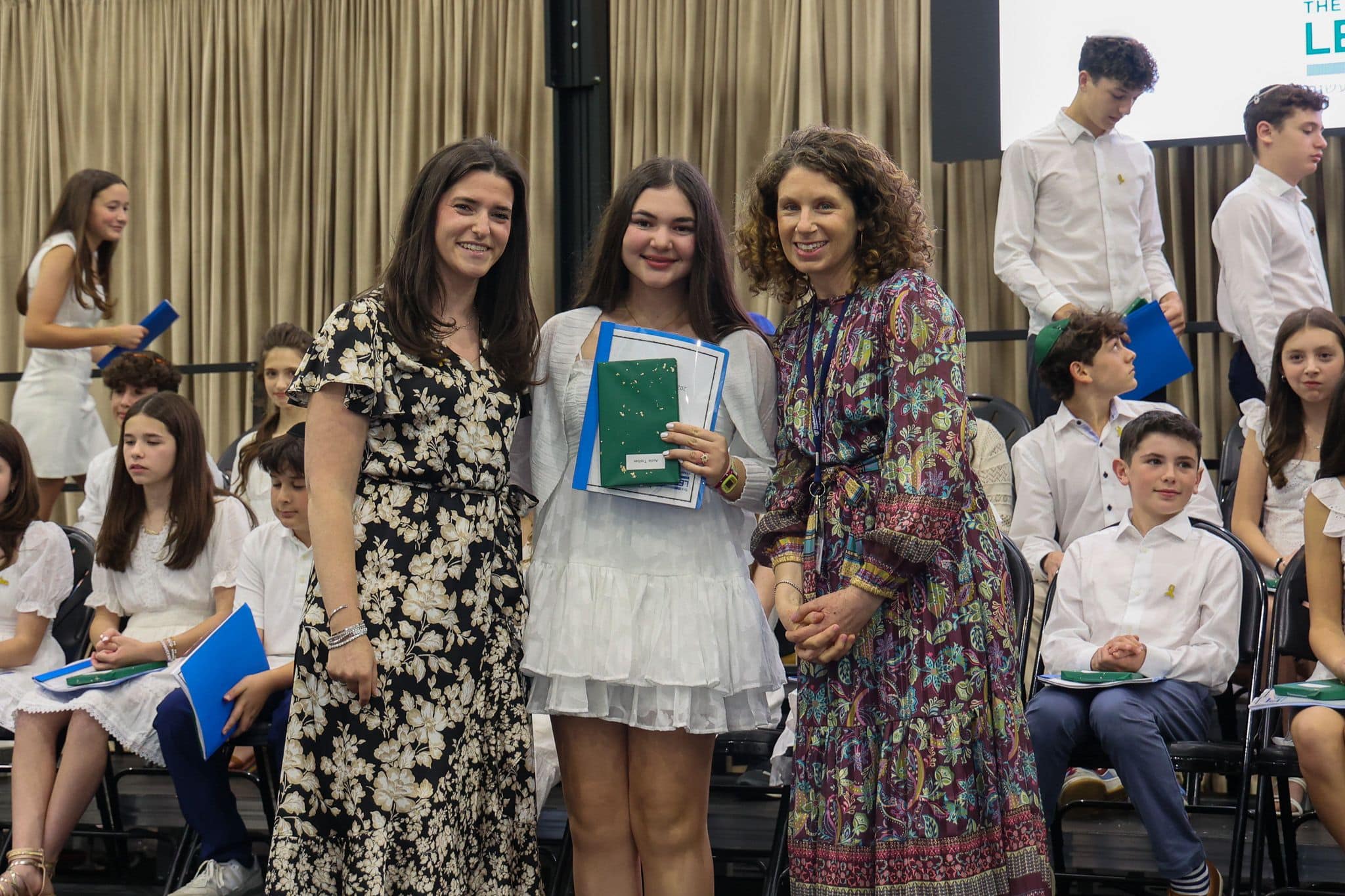 A group of three people, one in the middle and two on each side, are smiling for camera. They are at a ceremony, as shown by the backdrop displaying "The Leffell School." The central figure, a younger student in white clothes , on the sides are two women in floral dresses.