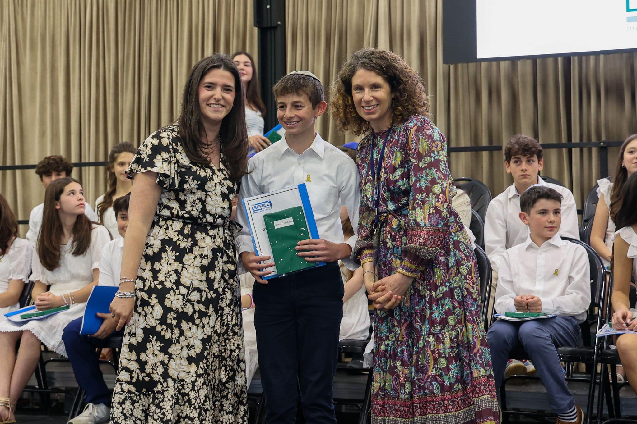 A group of three people, one in the middle and two on each side, are smiling for camera. They are at a ceremony, as shown by the backdrop displaying "The Leffell School." The central figure, a younger student in white clothes , on the sides are two women in floral dresses.