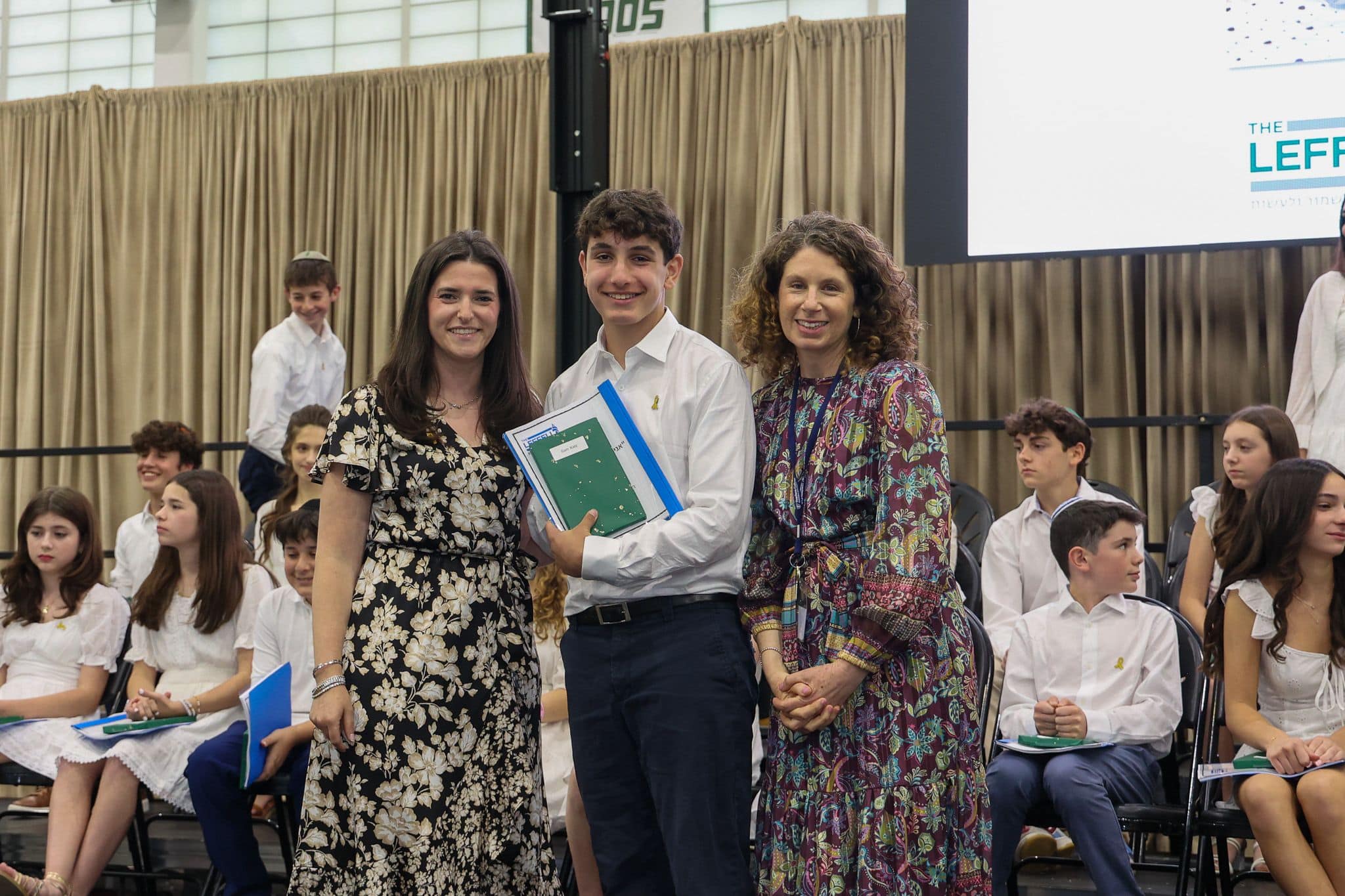 A group of three people, one in the middle and two on each side, are smiling for camera. They are at a ceremony, as shown by the backdrop displaying "The Leffell School." The central figure, a younger student in white clothes , on the sides are two women in floral dresses.