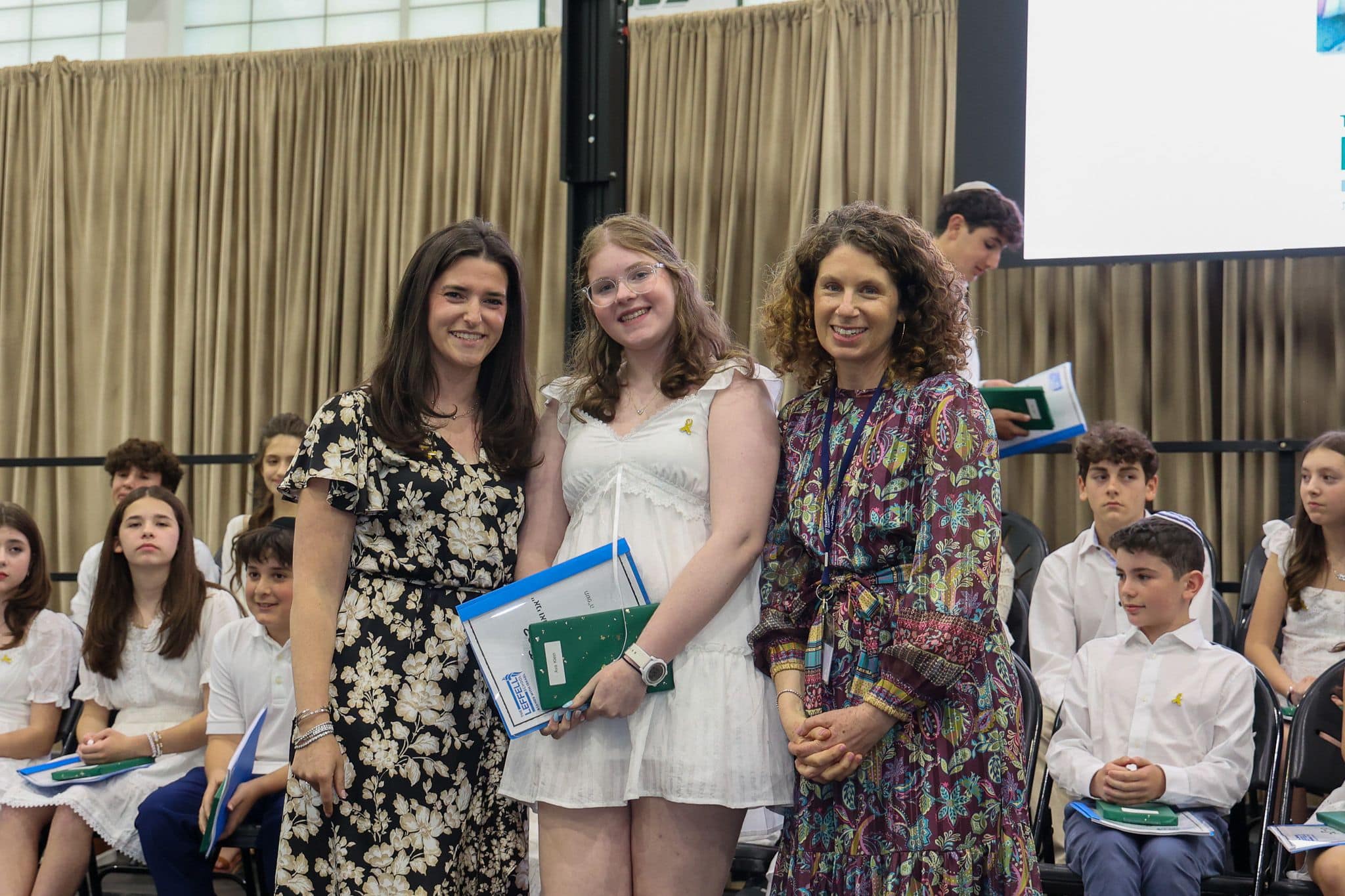 A group of three people, one in the middle and two on each side, are smiling for camera. They are at a ceremony, as shown by the backdrop displaying "The Leffell School." The central figure, a younger student in white clothes , on the sides are two women in floral dresses.