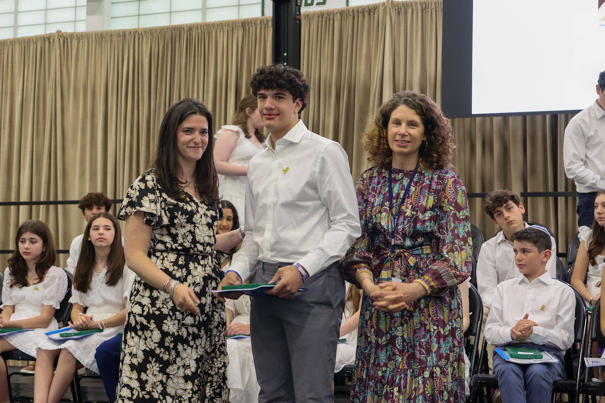 A group of three people, one in the middle and two on each side, are smiling for camera. They are at a ceremony, as shown by the backdrop displaying "The Leffell School." The central figure, a younger student in white clothes , on the sides are two women in floral dresses.