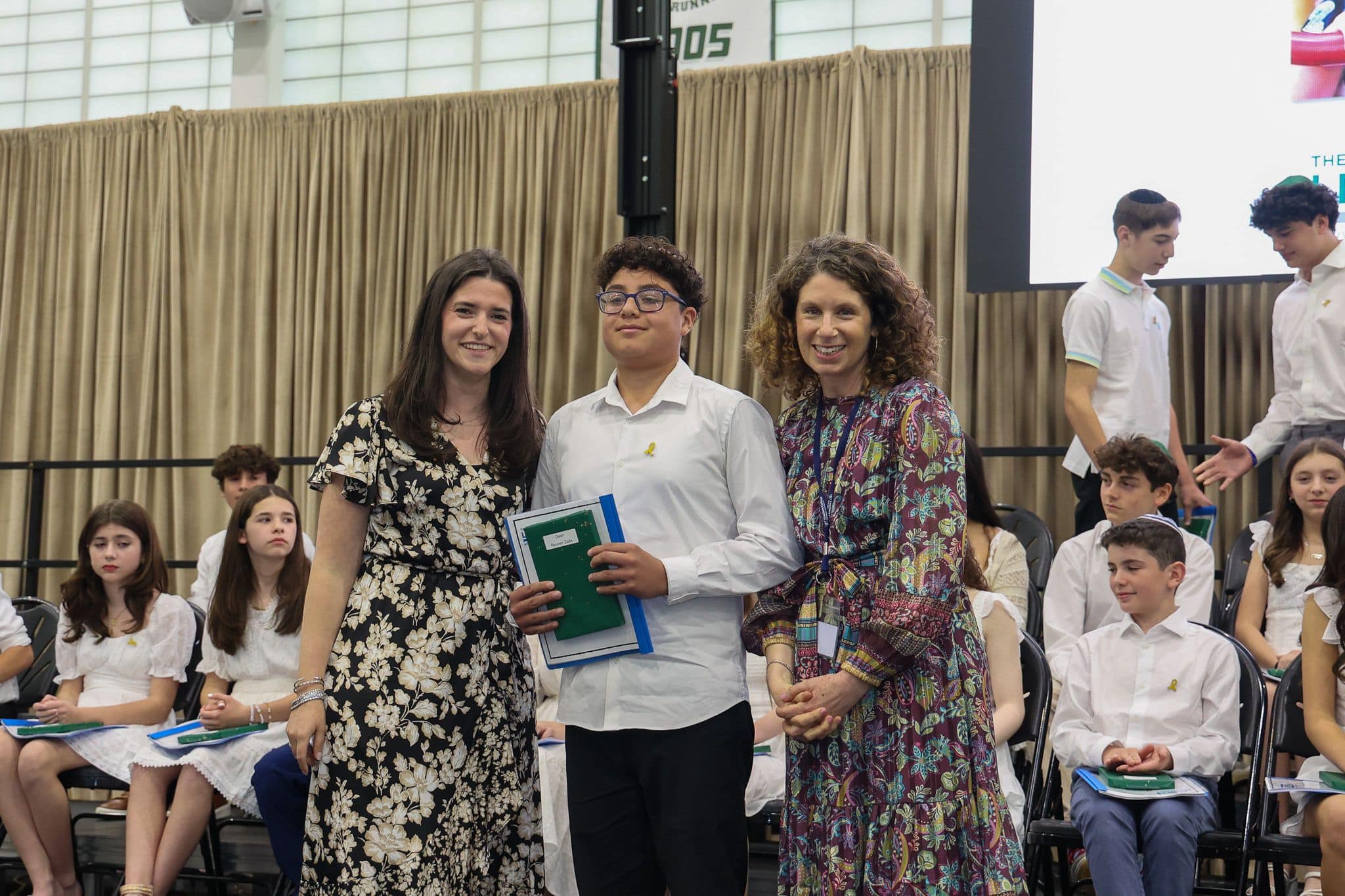 A group of three people, one in the middle and two on each side, are smiling for camera. They are at a ceremony, as shown by the backdrop displaying "The Leffell School." The central figure, a younger student in white clothes , on the sides are two women in floral dresses.