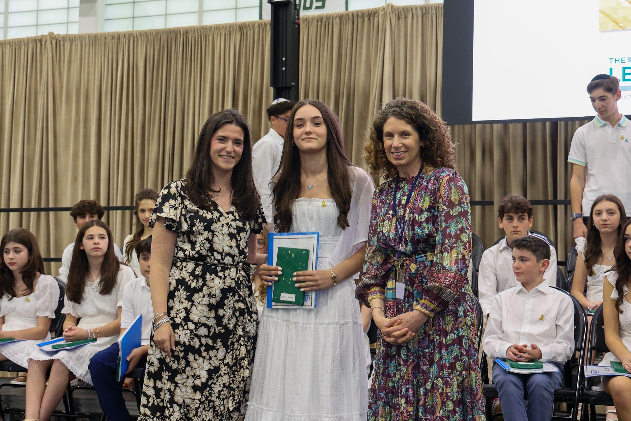 A group of three people, one in the middle and two on each side, are smiling for camera. They are at a ceremony, as shown by the backdrop displaying "The Leffell School." The central figure, a younger student in white clothes , on the sides are two women in floral dresses.
