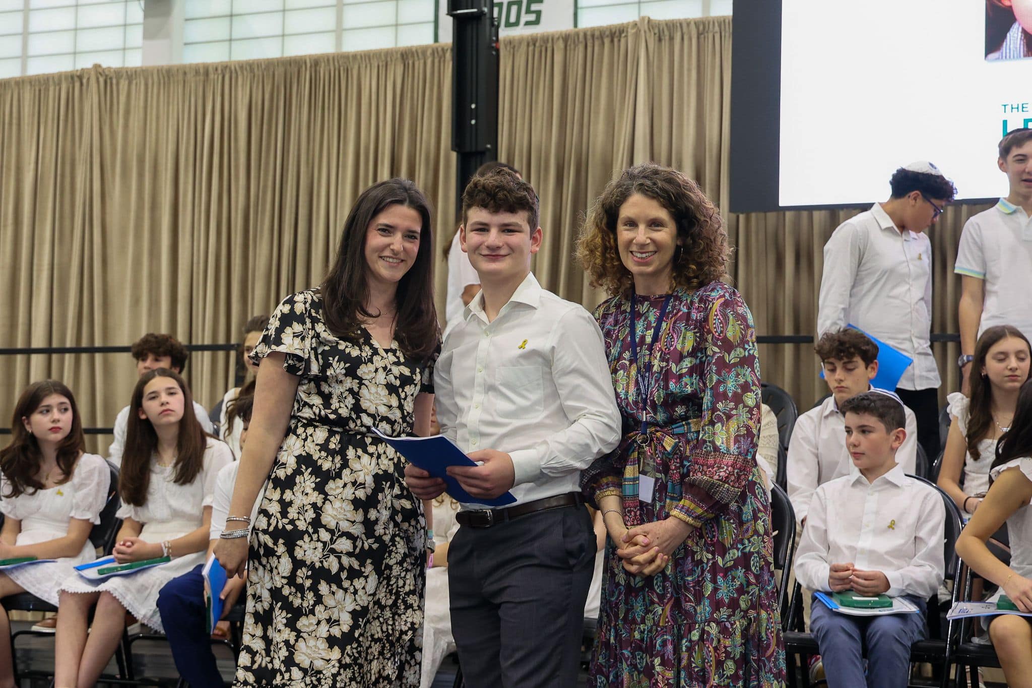 A group of three people, one in the middle and two on each side, are smiling for camera. They are at a ceremony, as shown by the backdrop displaying "The Leffell School." The central figure, a younger student in white clothes , on the sides are two women in floral dresses.