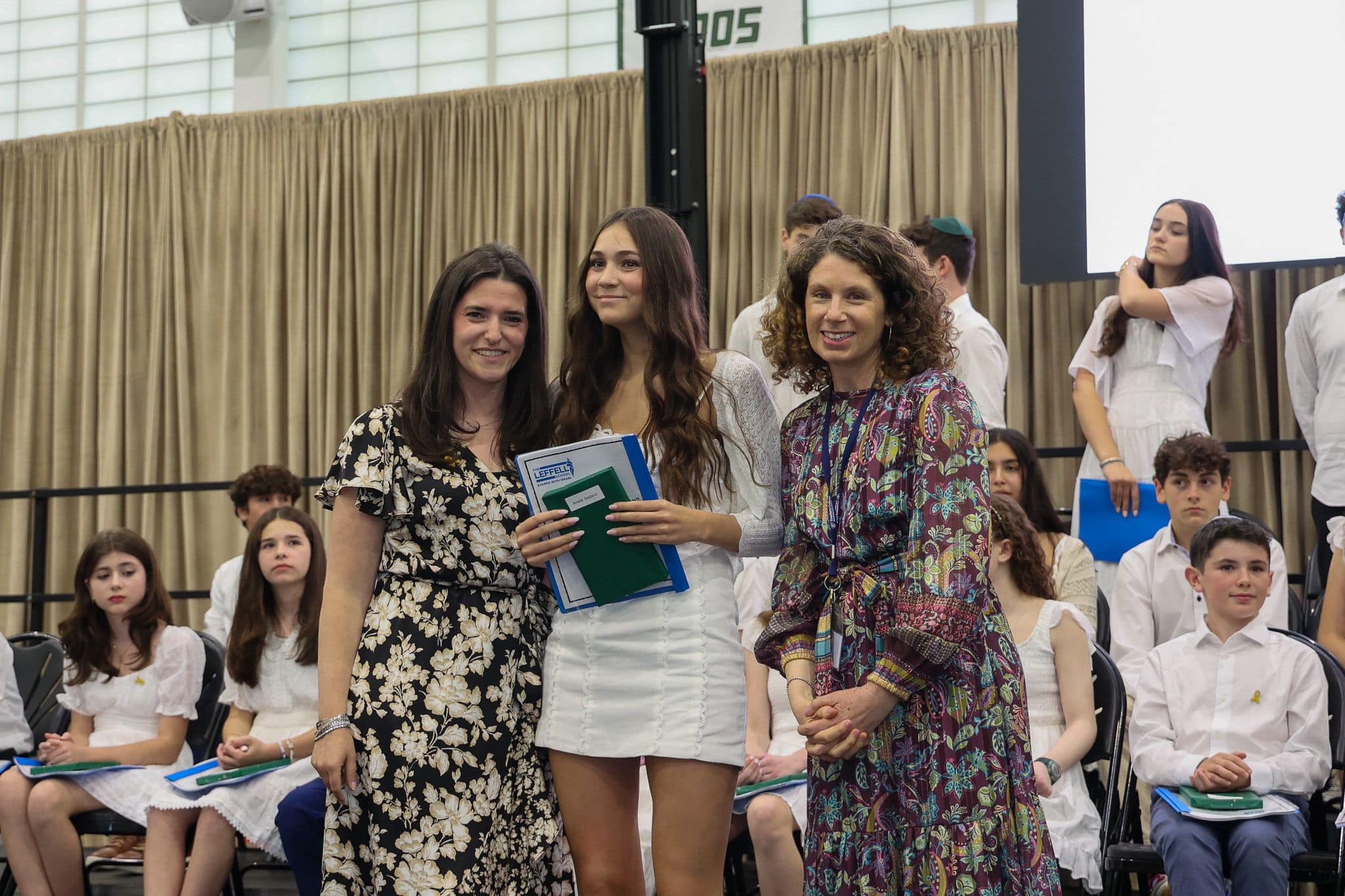 A group of three people, one in the middle and two on each side, are smiling for camera. They are at a ceremony, as shown by the backdrop displaying "The Leffell School." The central figure, a younger student in white clothes , on the sides are two women in floral dresses.