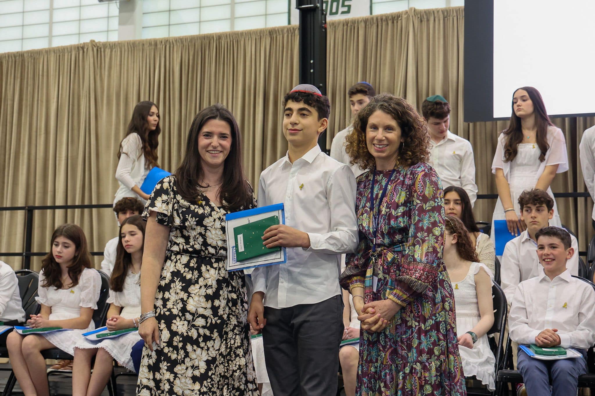 A group of three people, one in the middle and two on each side, are smiling for camera. They are at a ceremony, as shown by the backdrop displaying "The Leffell School." The central figure, a younger student in white clothes , on the sides are two women in floral dresses.