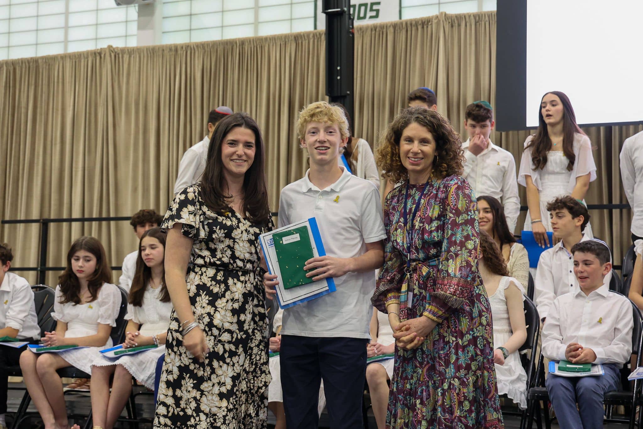 A group of three people, one in the middle and two on each side, are smiling for camera. They are at a ceremony, as shown by the backdrop displaying "The Leffell School." The central figure, a younger student in white clothes , on the sides are two women in floral dresses.
