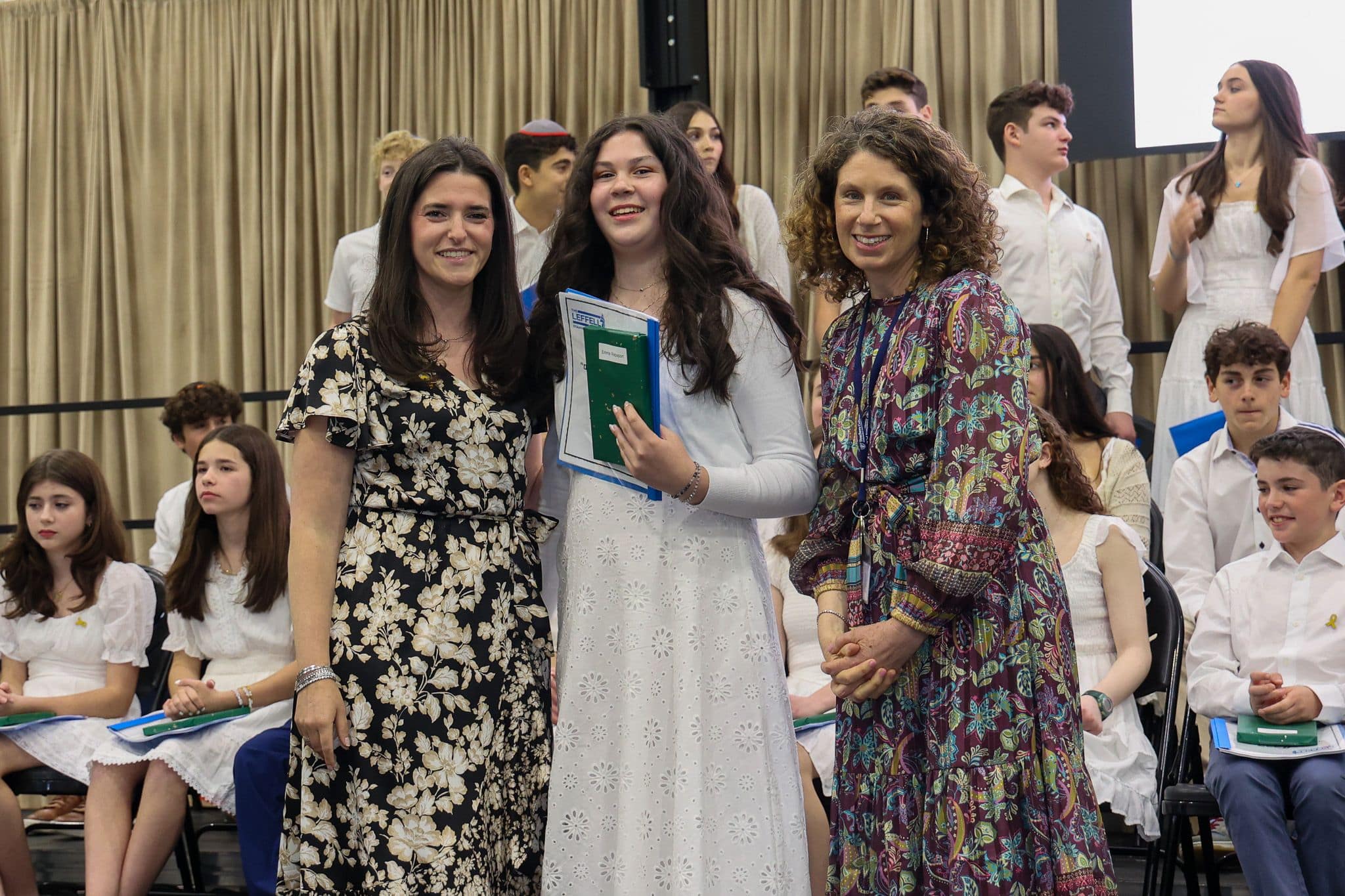A group of three people, one in the middle and two on each side, are smiling for camera. They are at a ceremony, as shown by the backdrop displaying "The Leffell School." The central figure, a younger student in white clothes , on the sides are two women in floral dresses.
