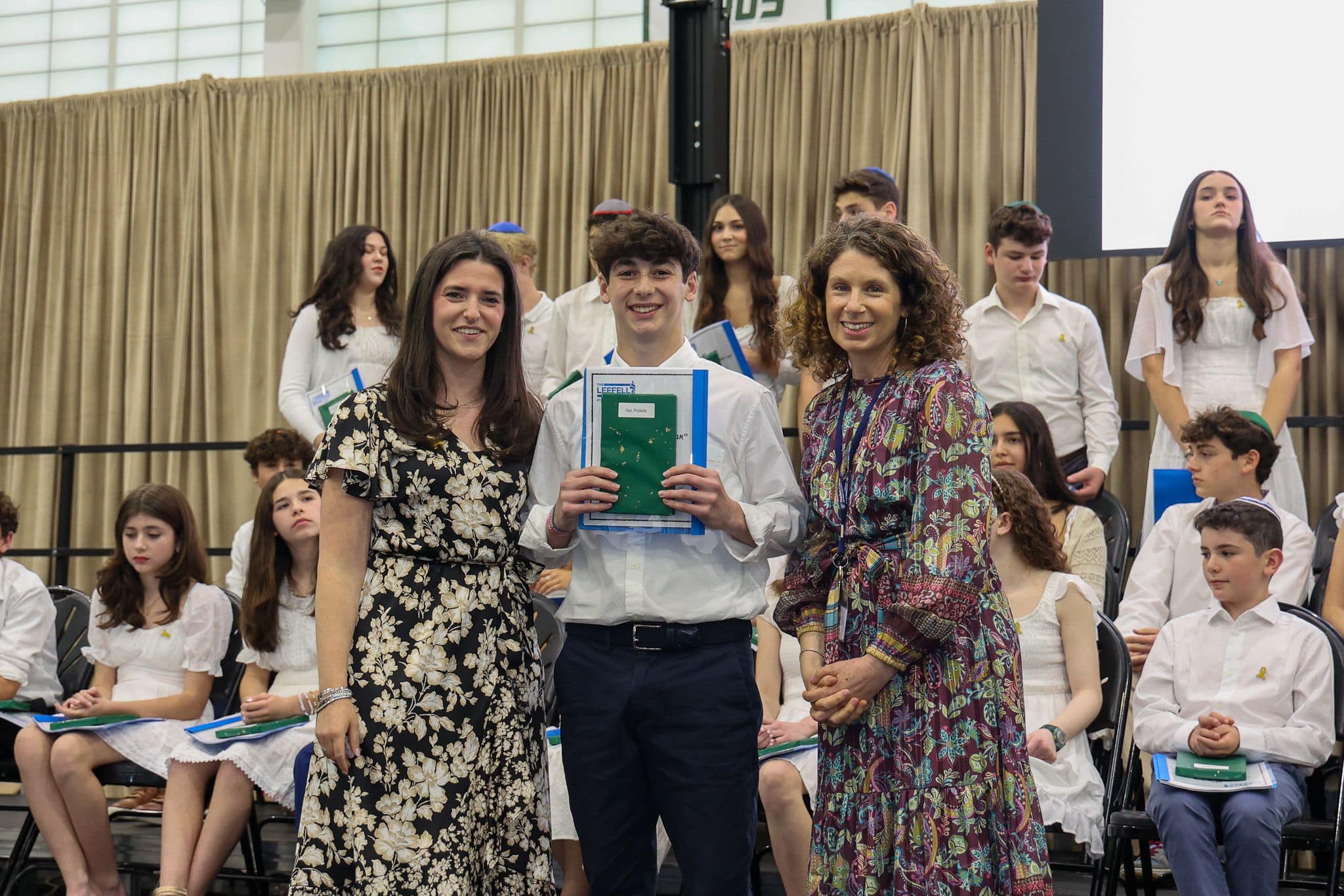 A group of three people, one in the middle and two on each side, are smiling for camera. They are at a ceremony, as shown by the backdrop displaying "The Leffell School." The central figure, a younger student in white clothes , on the sides are two women in floral dresses.