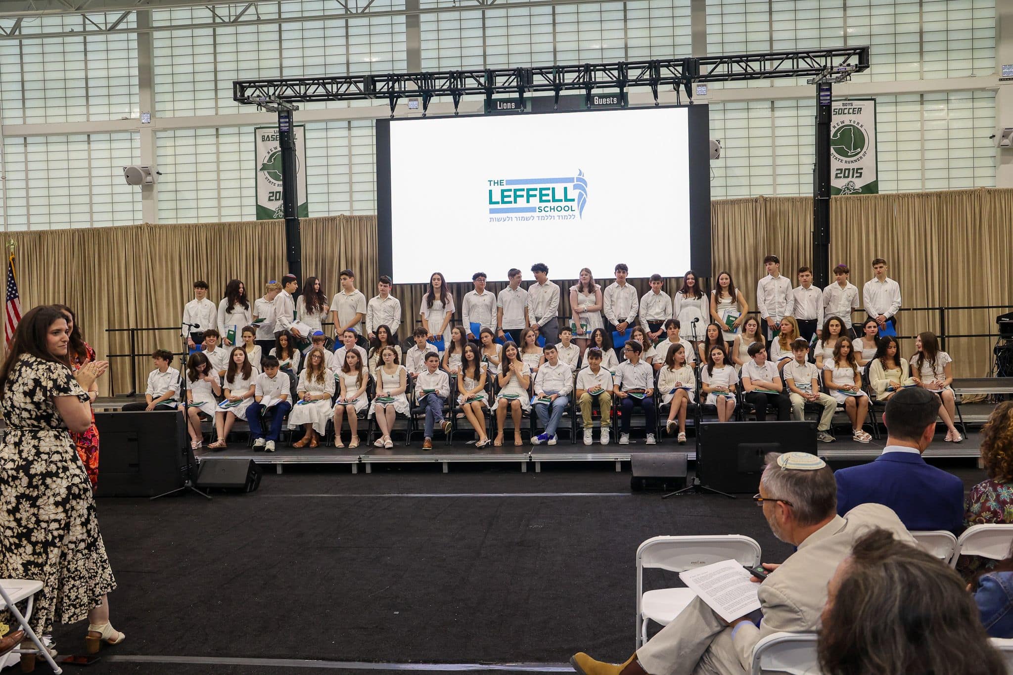 A group of students dressed in white sit on a stage with a large screen behind them displaying the leffel school logo