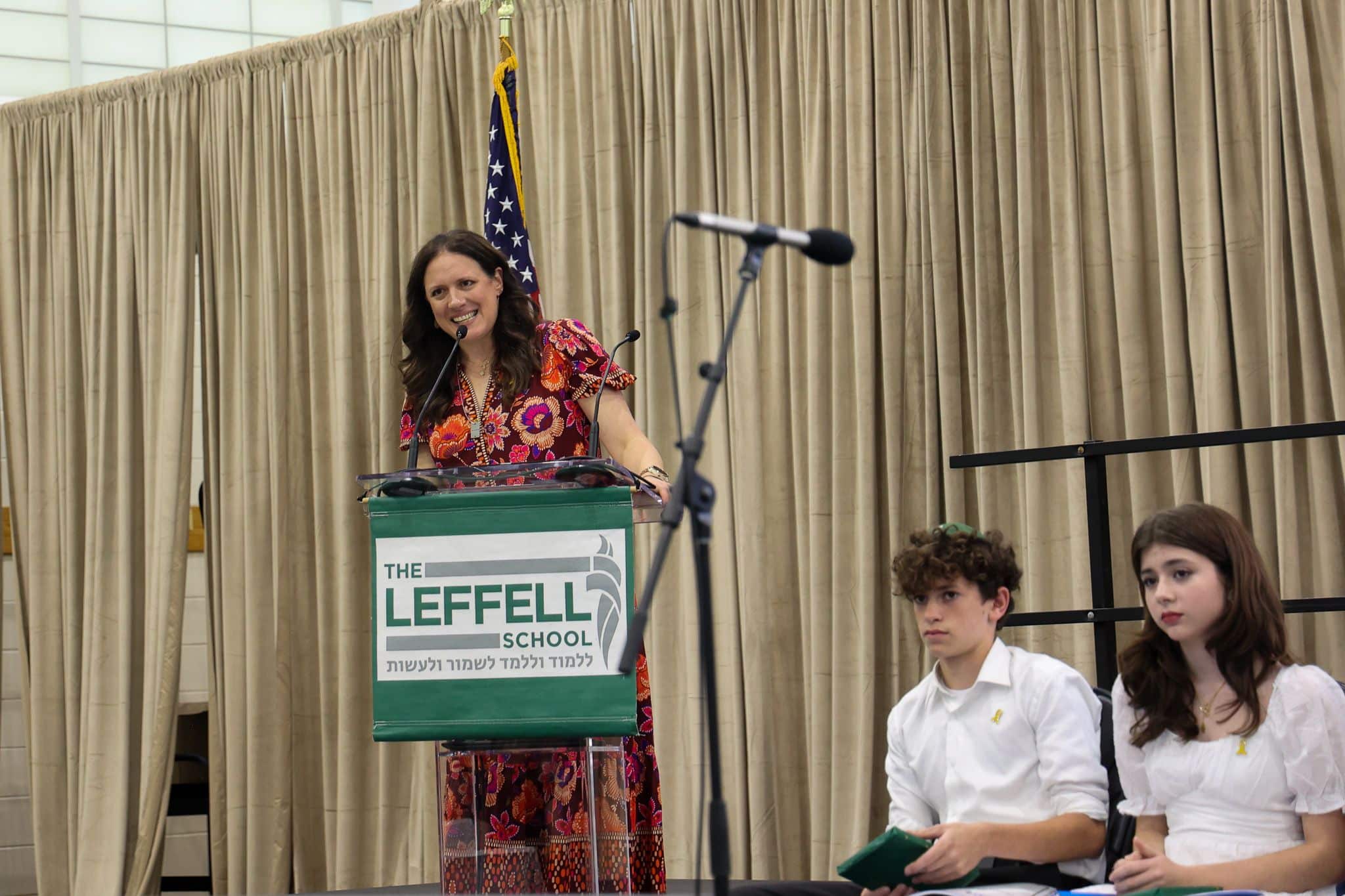 A woman speaks at a podium labeled "The Leffell School," with an American flag on the left. She wears a red outfit, set against beige curtains.