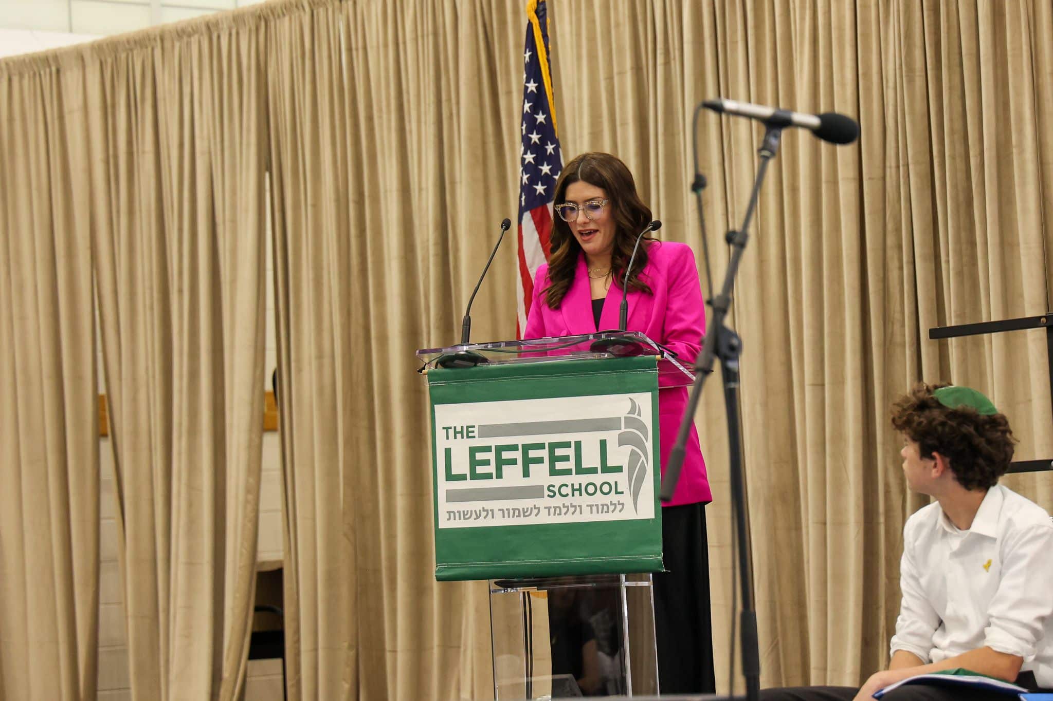 A woman speaks at a podium labeled "The Leffell School," with an American flag on the left. She wears a pink blazer, set against beige curtains.