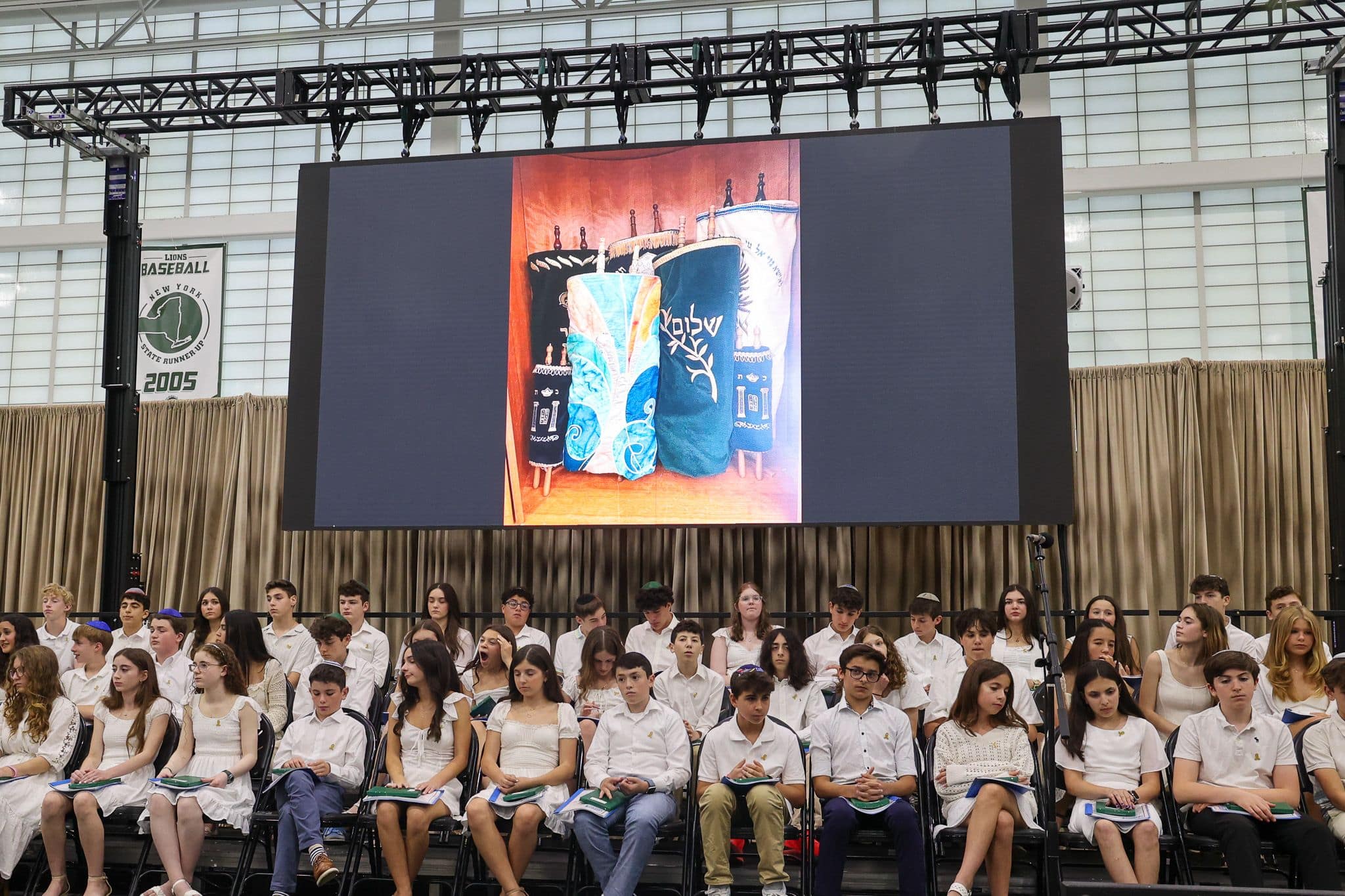 A group of students dressed in white sit on a stage with a large screen behind them displaying the leffel school logo