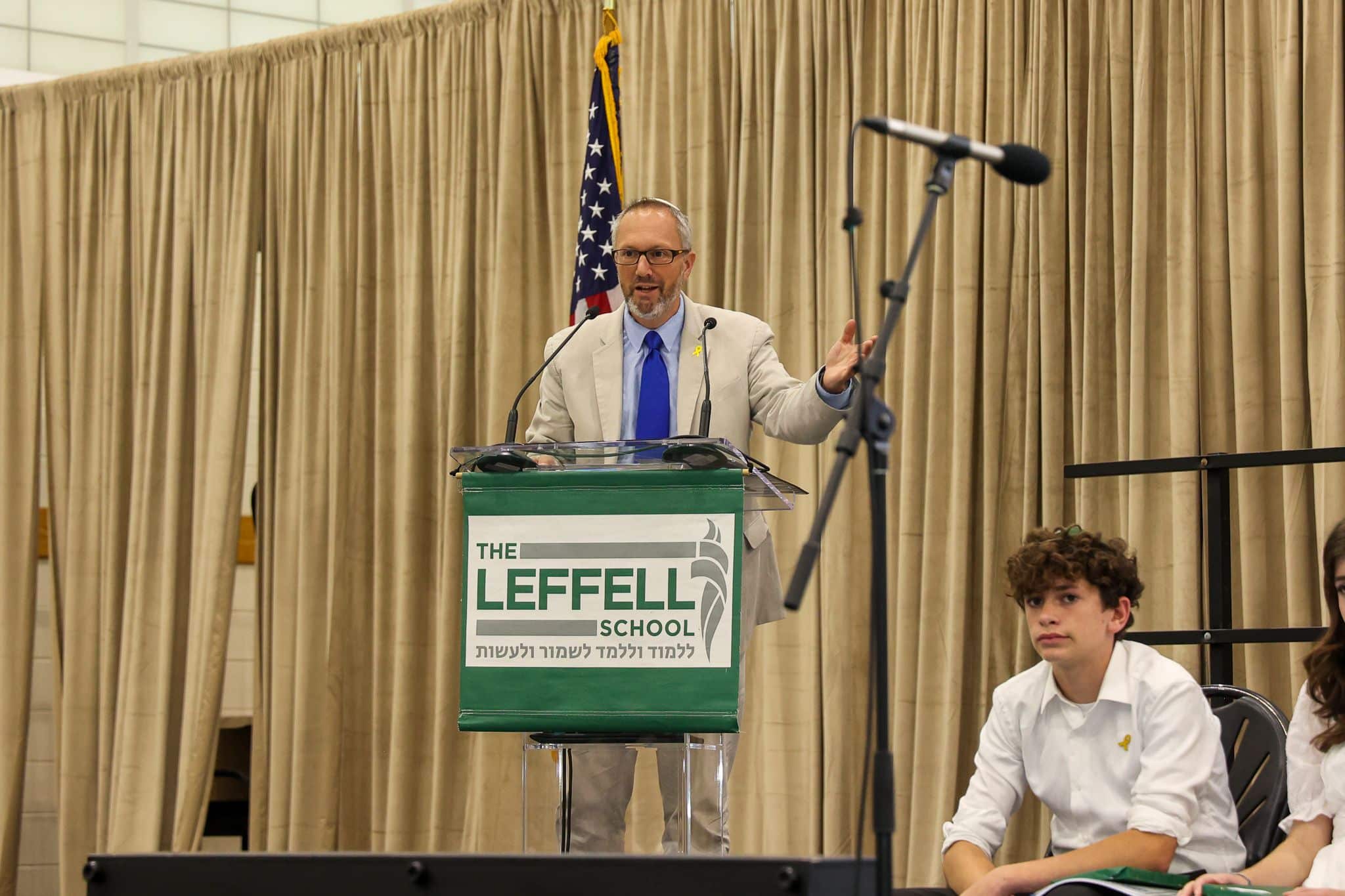 A man speaks at a podium labeled "The Leffell School," with an American flag on the left. He wears a tan suit , set against beige curtains.