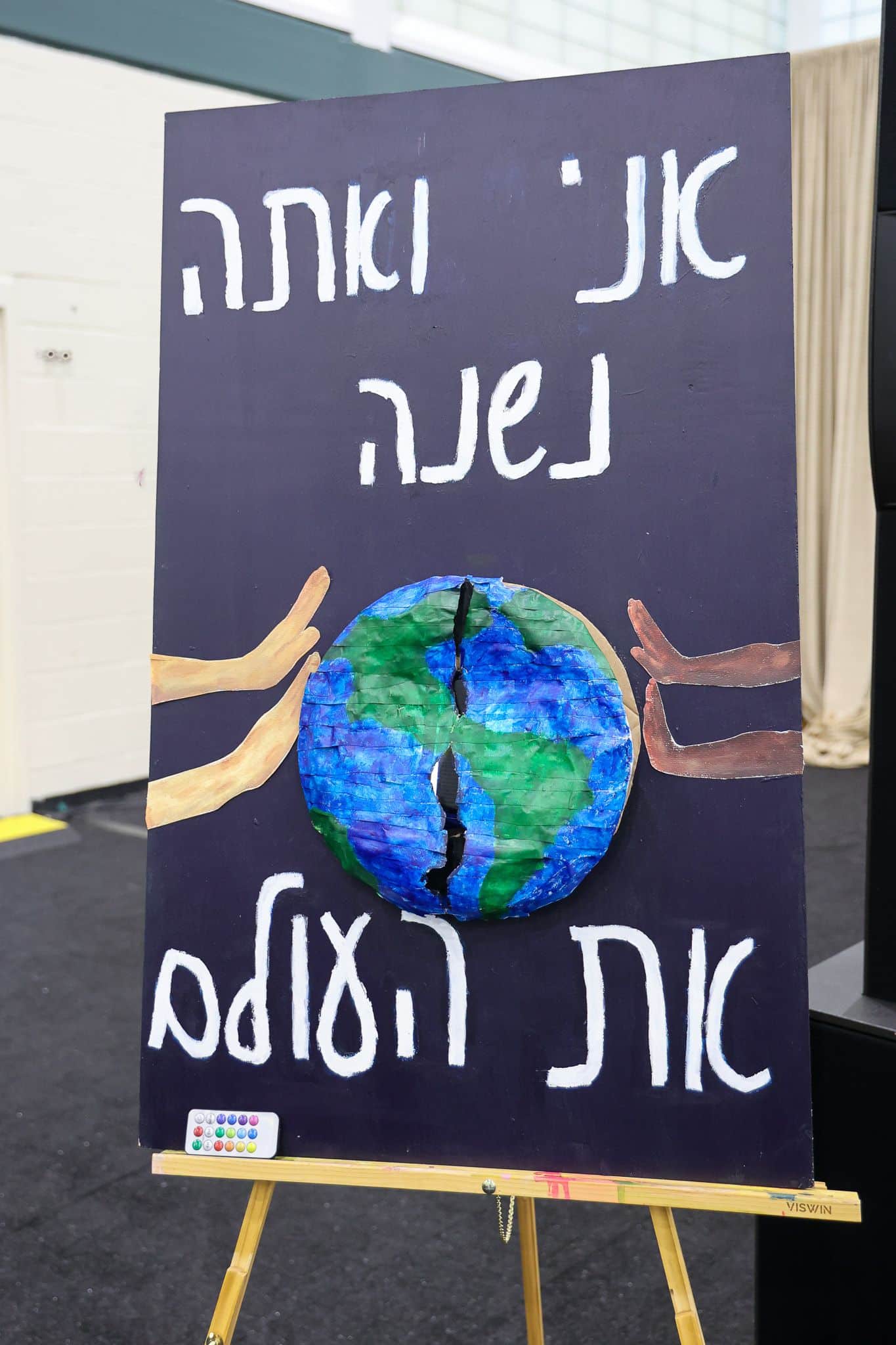 A decorated easel displays a Earth-themed artwork with Hebrew text in a gym or hall. Nearby, a colorful banner and tables with green items and tablecloths.