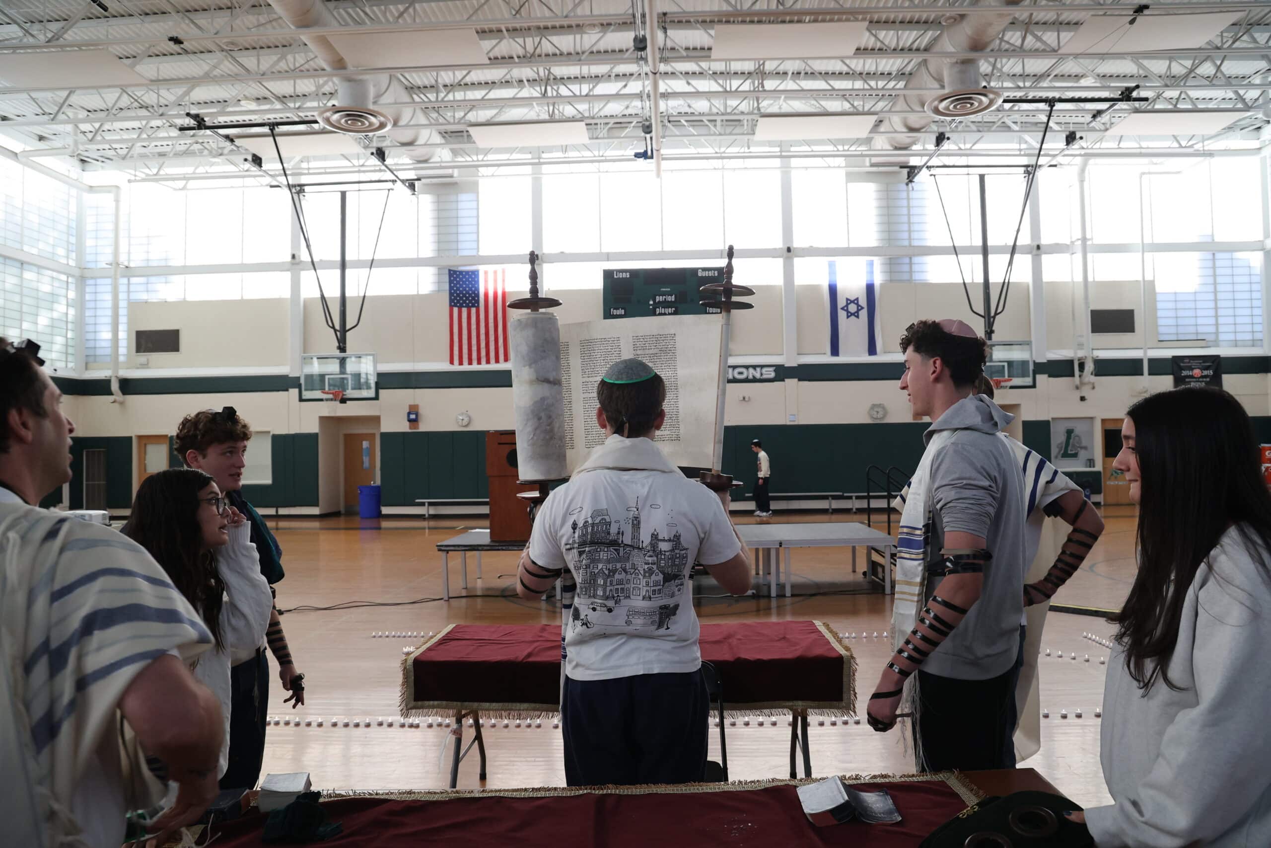 Teenage student hold Torah aloft revealing the Hebrew words on the scroll. He is surrounded by peers and standing in a gym with the American and Israeli flags on the wall.