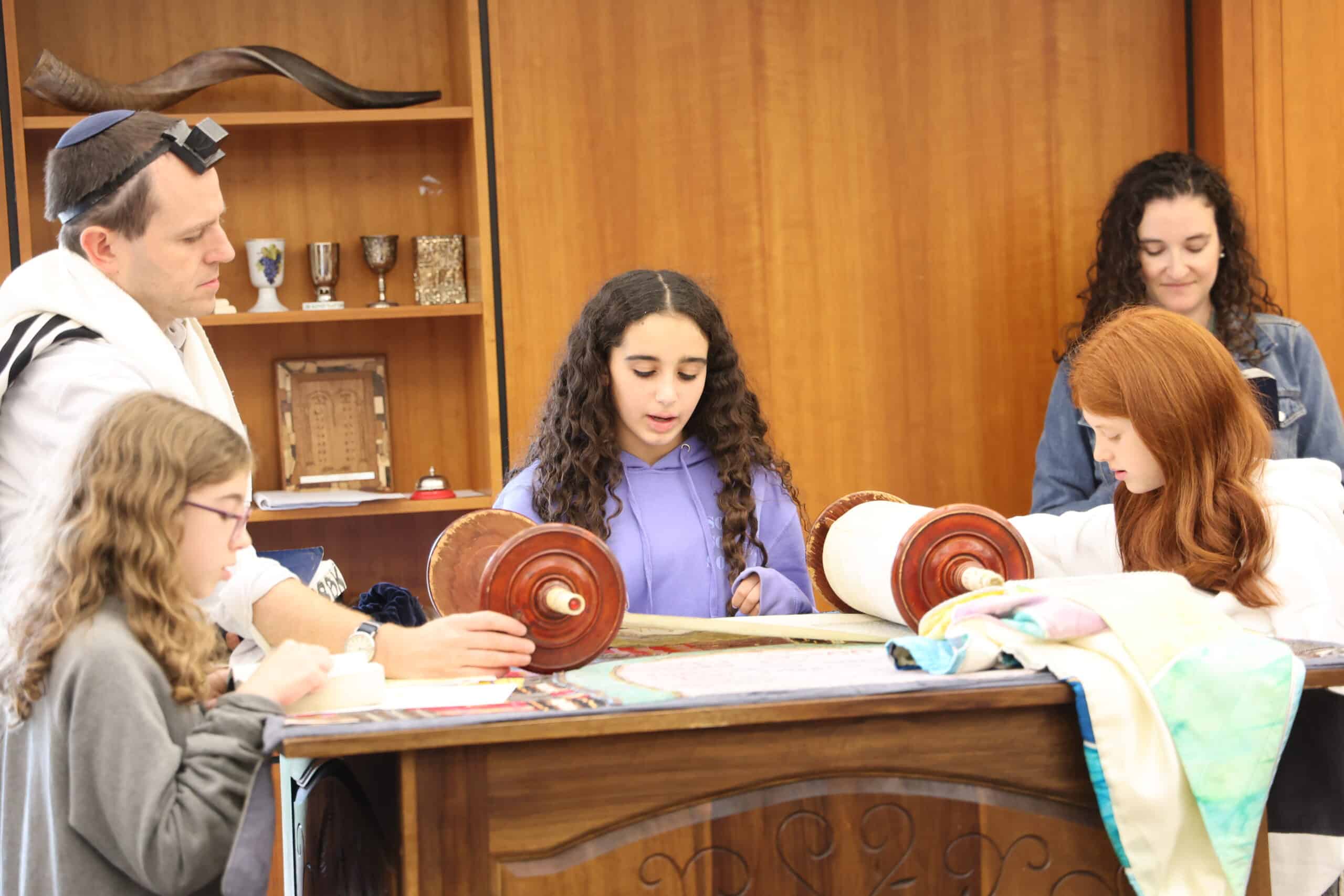 Female student stands at a bimah reading from the Torah. Two students and two teachers stand around her. A shelf of Judaica is in the background on a wooden shelf